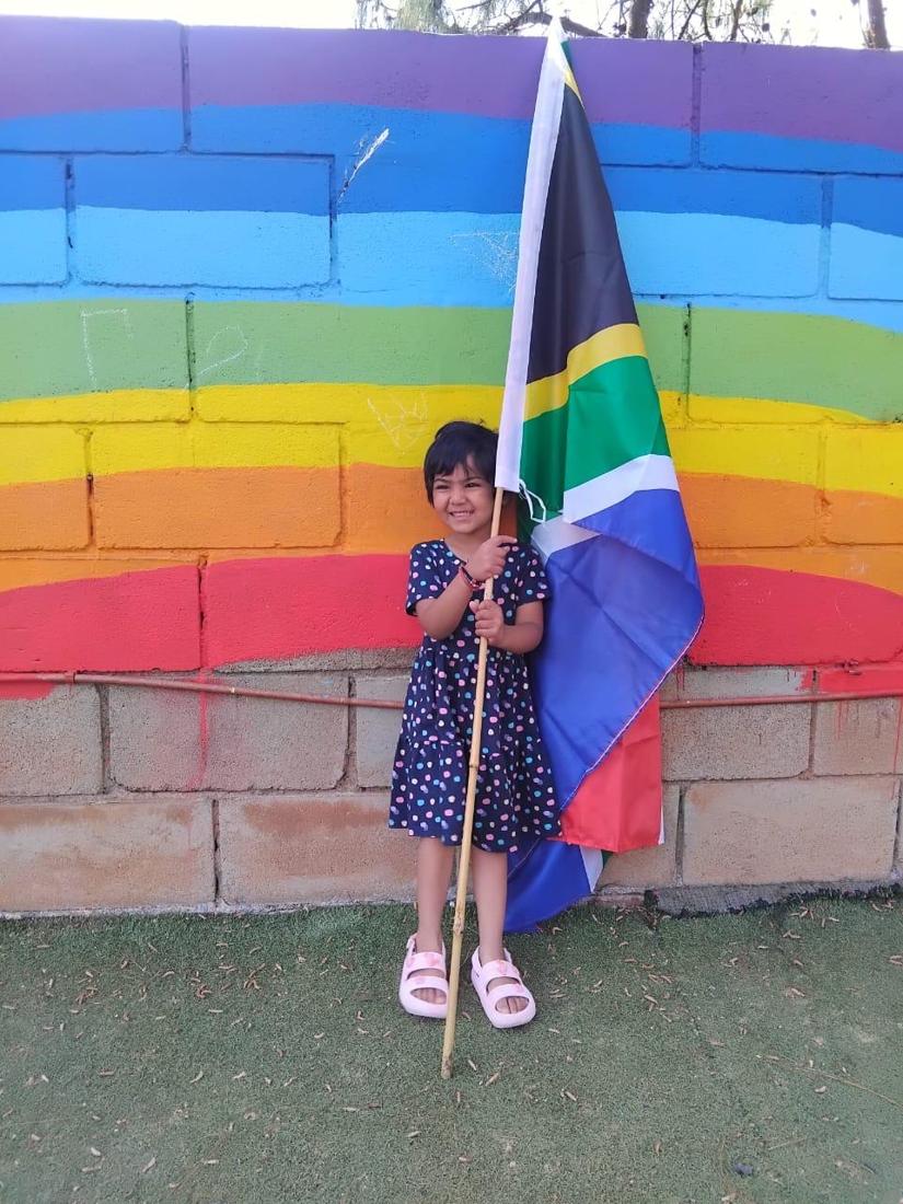 A smiling girl holding the South African flag in front of the rainbow wall on Heritage Day